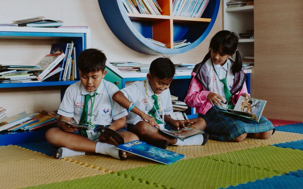 Three children in school uniforms reading books together in a vibrant library setting.