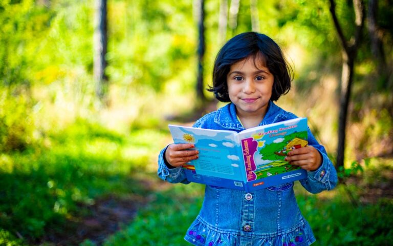 A young girl enjoys reading outdoors, holding a colorful book in a sunny park setting.