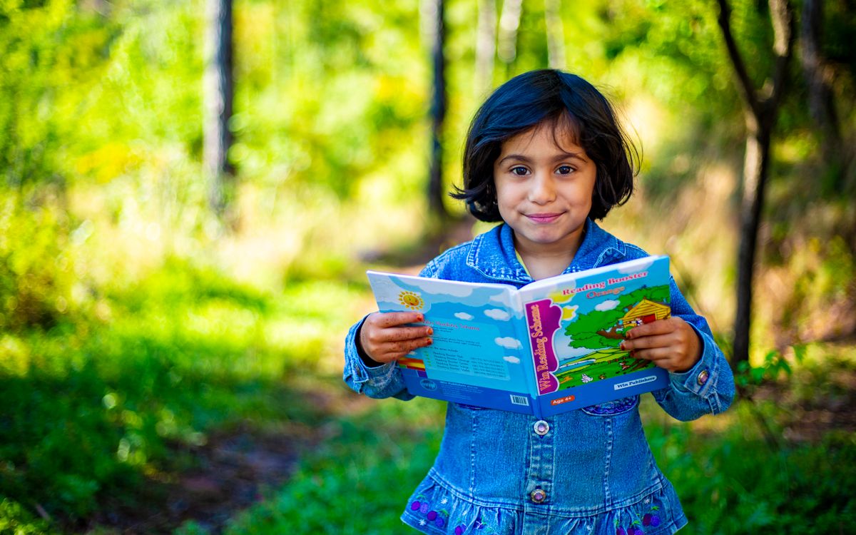 A young girl enjoys reading outdoors, holding a colorful book in a sunny park setting.