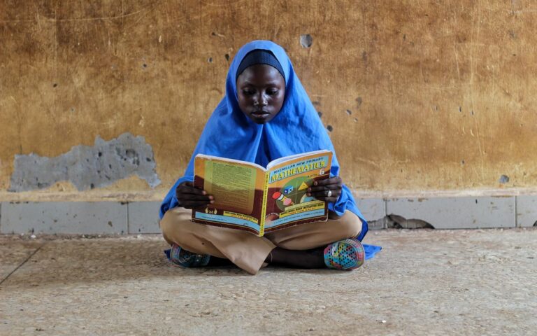 A young girl in hijab sitting on a floor reading a mathematics book intensely.