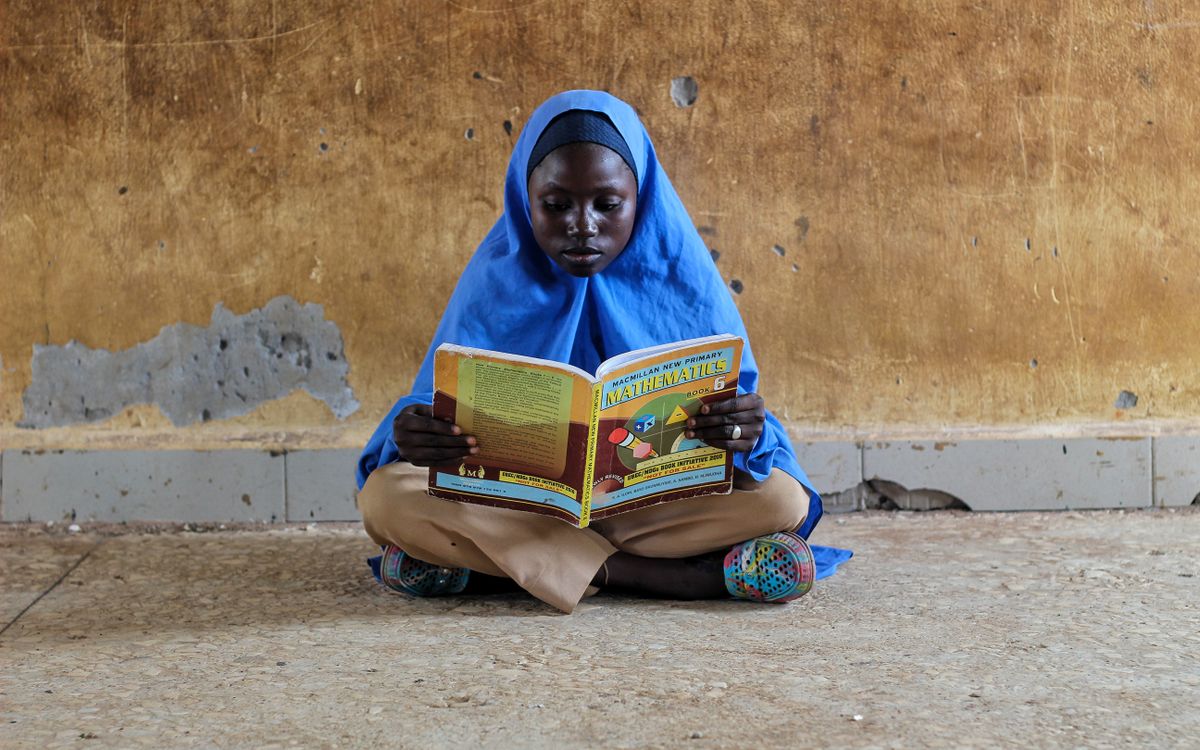 A young girl in hijab sitting on a floor reading a mathematics book intensely.