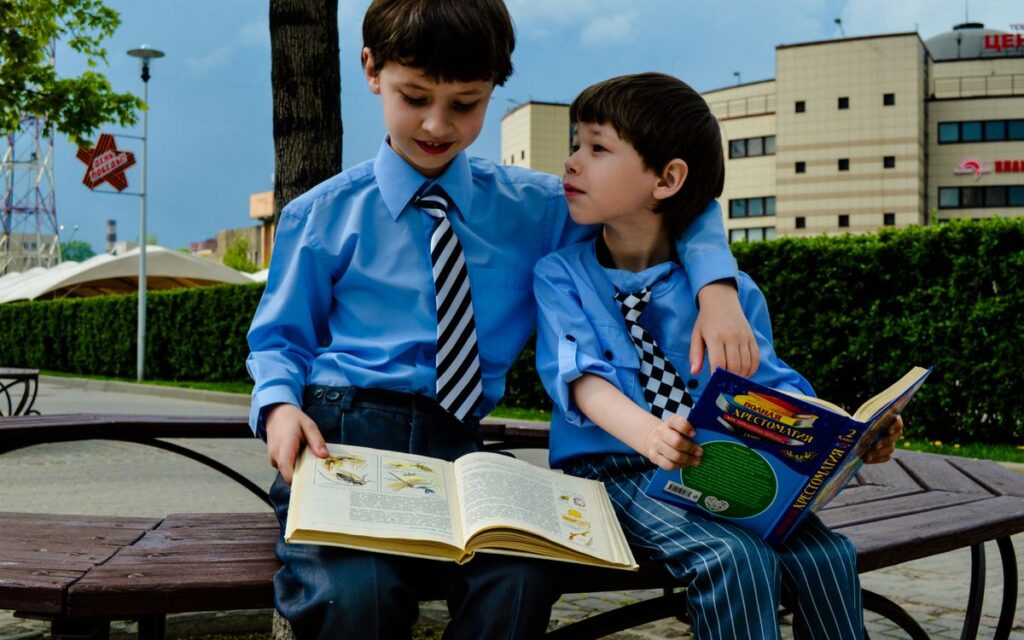 Two young boys in ties sit on a bench reading books in a sunny urban park.