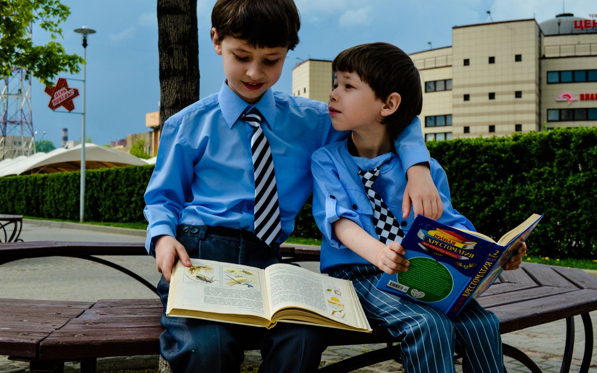 Two young boys in ties sit on a bench reading books in a sunny urban park.