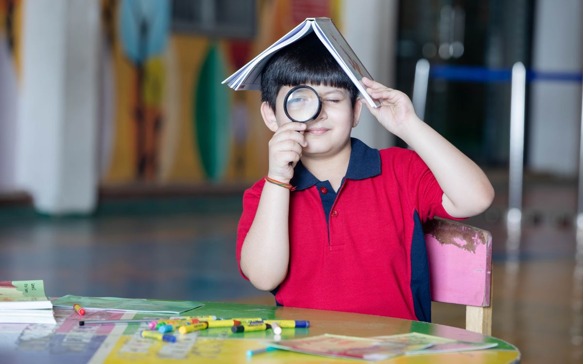 Child with book on head using magnifying glass at school desk.