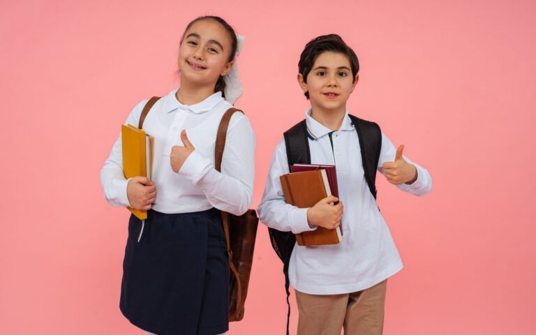 Two smiling schoolchildren give thumbs up with books and backpacks against a vibrant pink background.