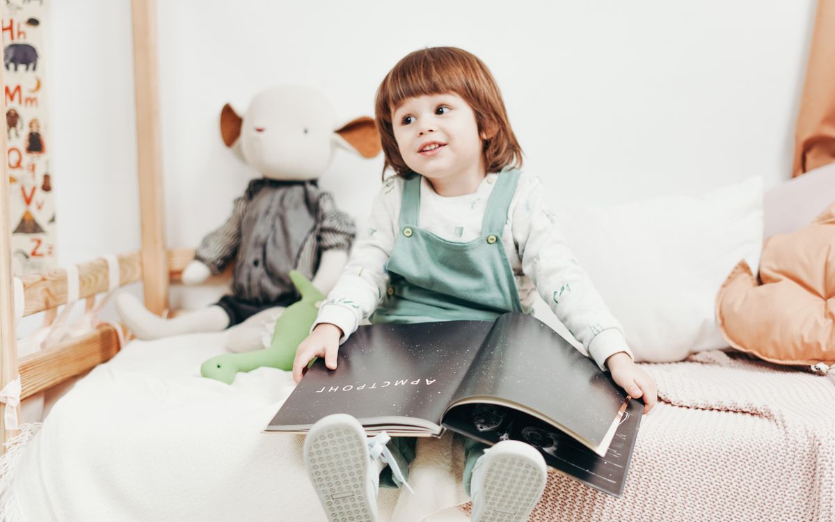 Adorable child sitting on a bed, enjoying a book in a bright playroom with toys.