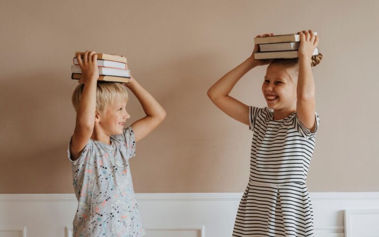 Two smiling children balancing books on their heads indoors, symbolizing fun in learning.