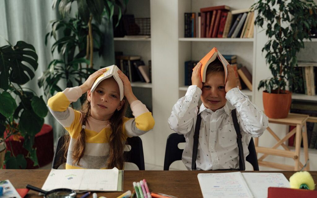 Two children playfully place books on their heads during a study session indoors.