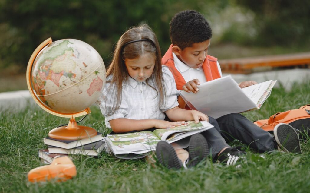 Two children sitting on grass reading books with a globe, promoting education.