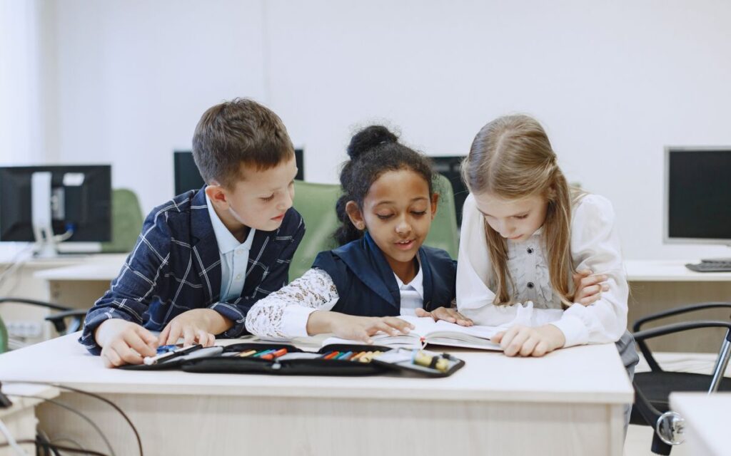 Three children working together at a desk in a classroom, focusing on a book.