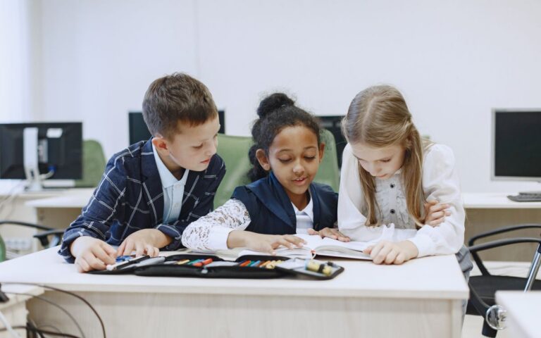 Three children working together at a desk in a classroom, focusing on a book.