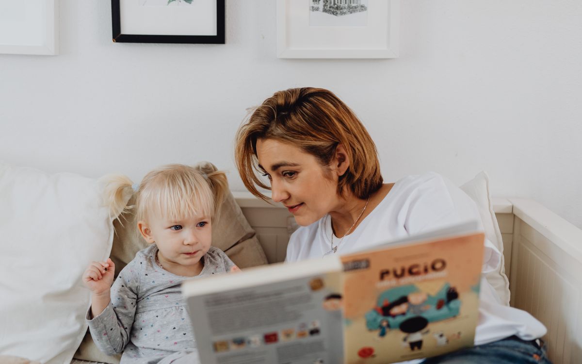 A mother and her daughter sharing a bonding moment while reading a children's book in the bedroom.
