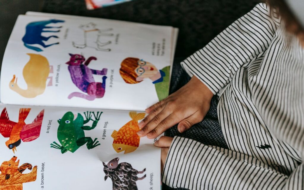 From above of crop anonymous child reading interesting book with colorful pictures of different animals