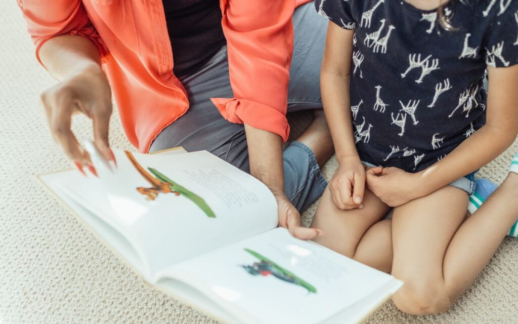 A mother and daughter sit on the floor, engaged in reading a colorful children's book.