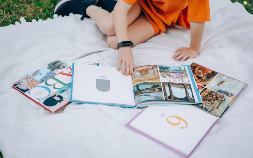 Young girl in orange dress reading books outdoors on a picnic blanket.