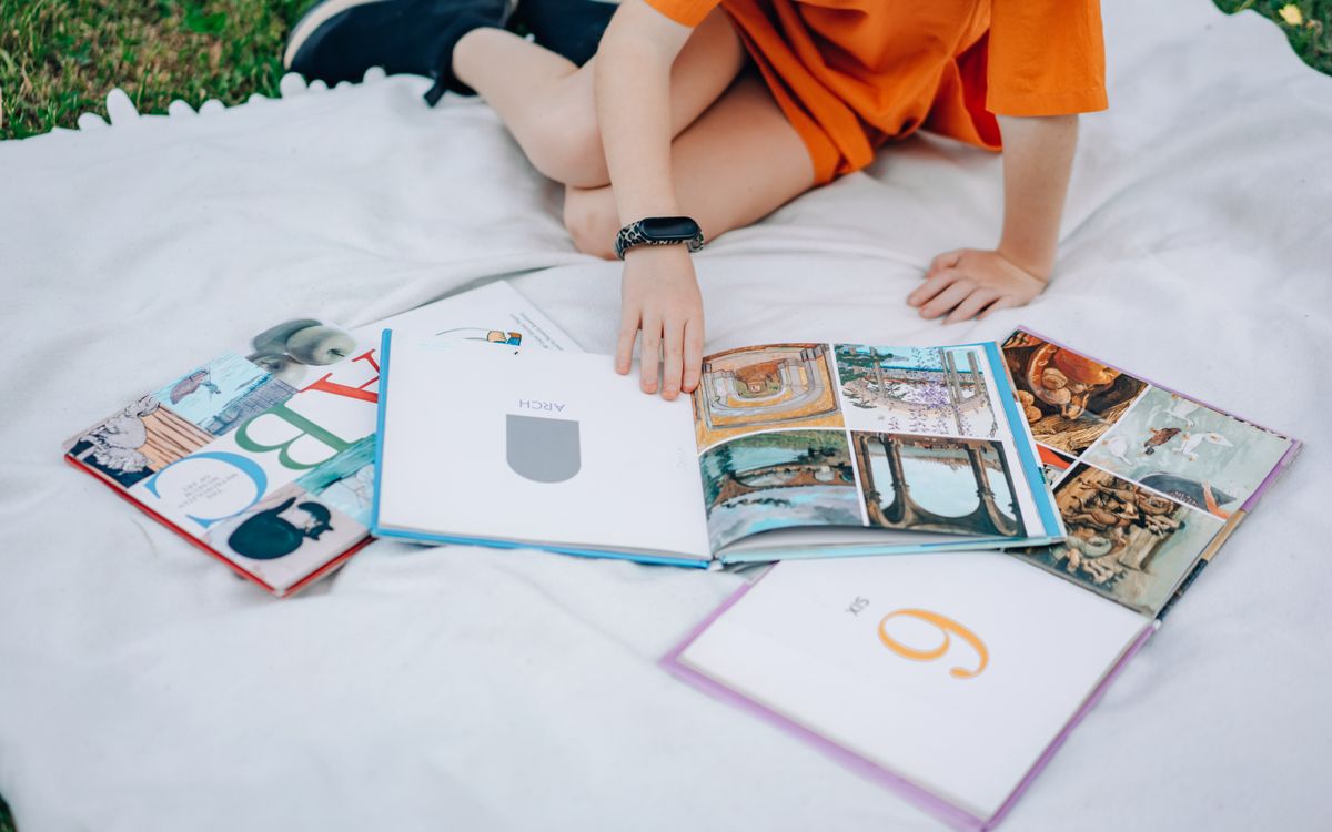 Young girl in orange dress reading books outdoors on a picnic blanket.