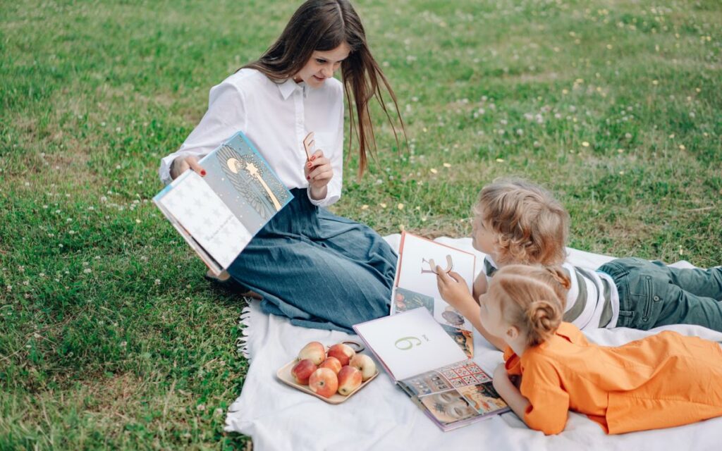 A teacher and two kids enjoy reading books on a picnic in a grassy park setting.