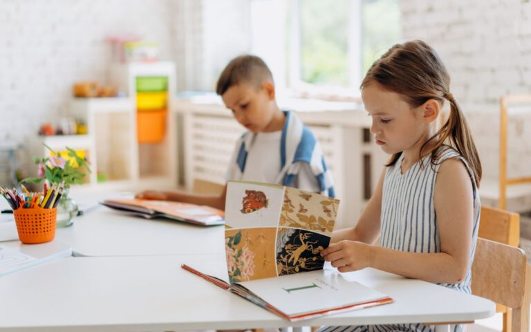 Two children reading books at a bright indoor classroom table filled with colorful supplies.