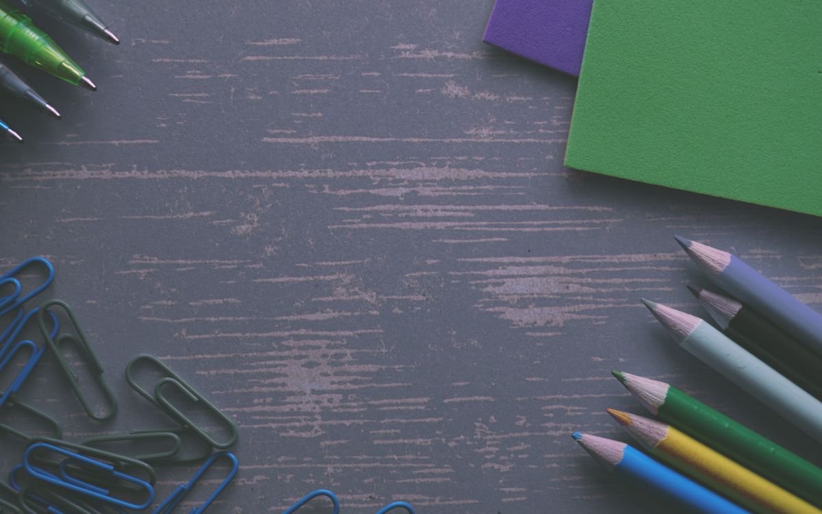 A flat lay of colored pencils, paper clips, and paper on a wooden desk.