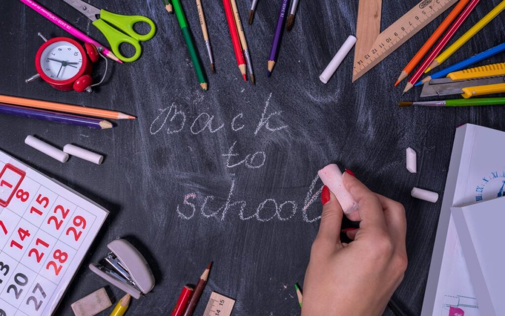 Hand writing 'back to school' on chalkboard surrounded by school supplies.
