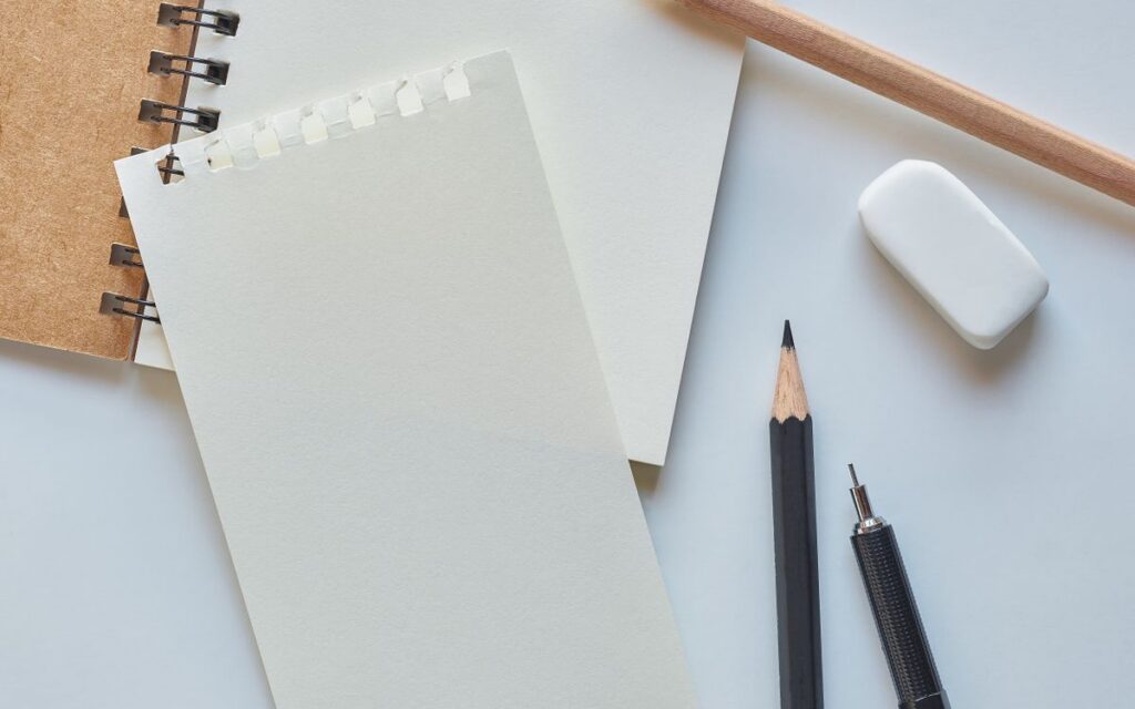 Flat lay of a spiral notebook, pencils, pen, and eraser on a white background.