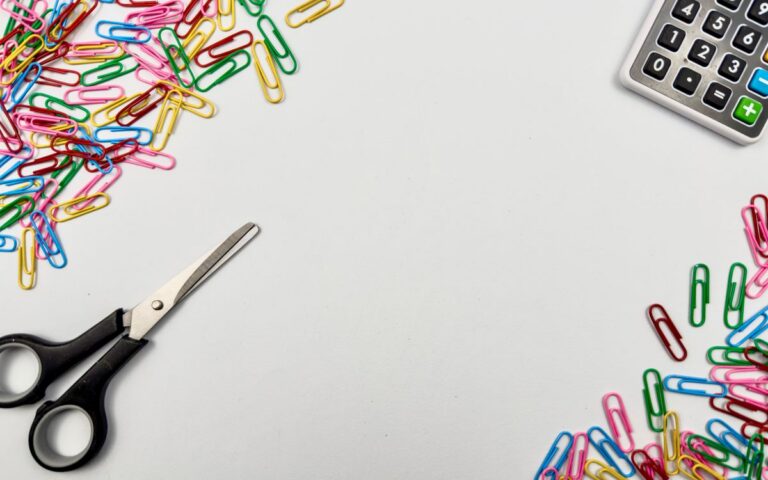 Brightly colored paperclips scattered on a desk with scissors and a calculator for a creative workspace vibe.