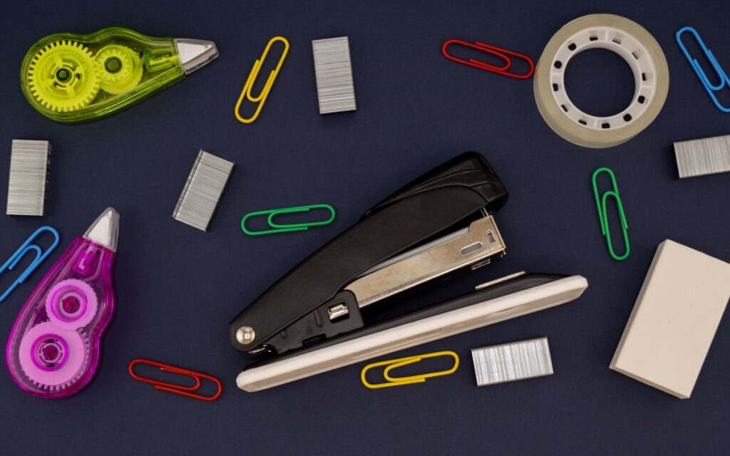 Flat lay of assorted office supplies including stapler, paper clips, and tape on dark background.