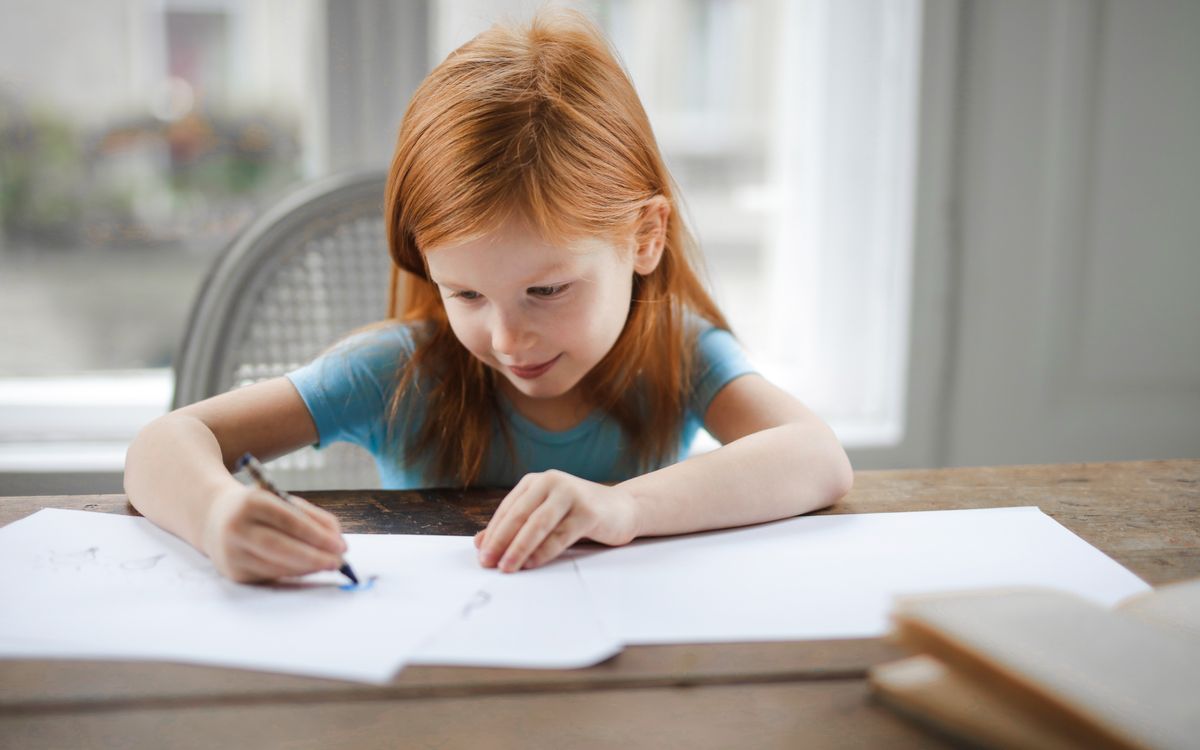 High angle of delighted redhead schoolgirl in casual clothes focusing and drawing with pencil on paper while sitting at