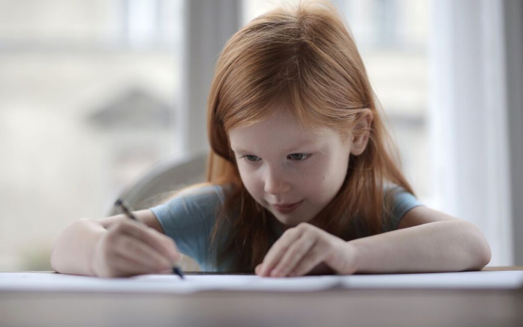 Adorable positive preschool child with red hair in casual wear sitting at table and drawing with pencil on paper sheet a