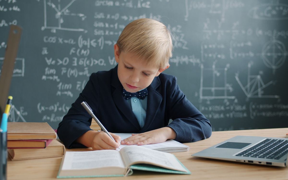 A young student diligently works on an assignment in a classroom with a laptop nearby.