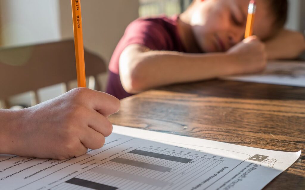 Crop unrecognizable schoolkid with pencil in hand writing on paper sheet with diagrams while sitting at table with broth
