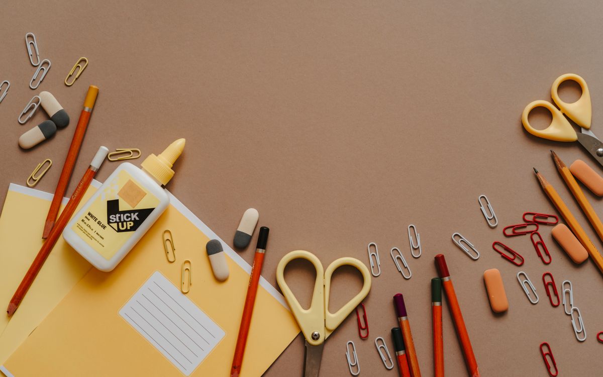 Flat lay of assorted stationery items like scissors, pencils, and paper clips on a brown background.