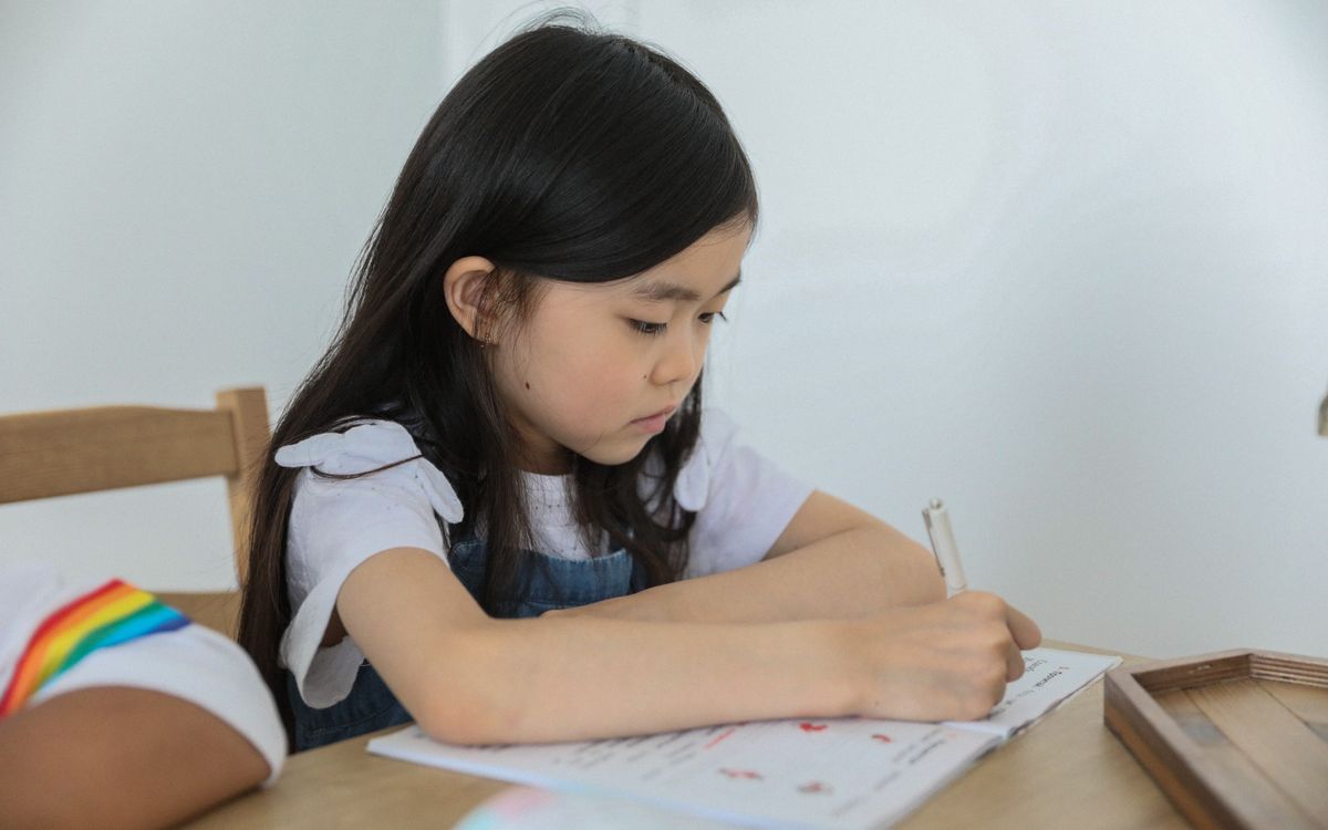 Concentrated ethnic schoolkid writing in activity book with pen while studying at table near unrecognizable black friend