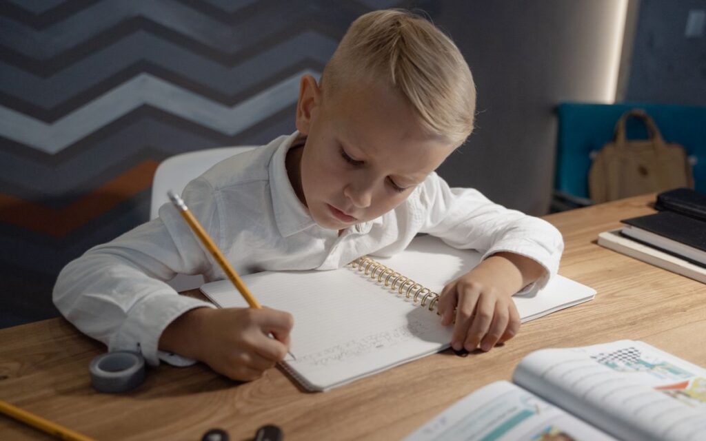 A young boy diligently writing homework in a notebook at a wooden desk indoors.