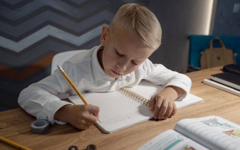 A young boy diligently writing homework in a notebook at a wooden desk indoors.