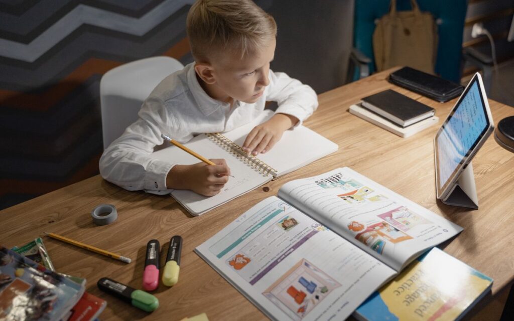 A focused young boy engages in distance learning with a tablet, books, and notebooks on a desk.