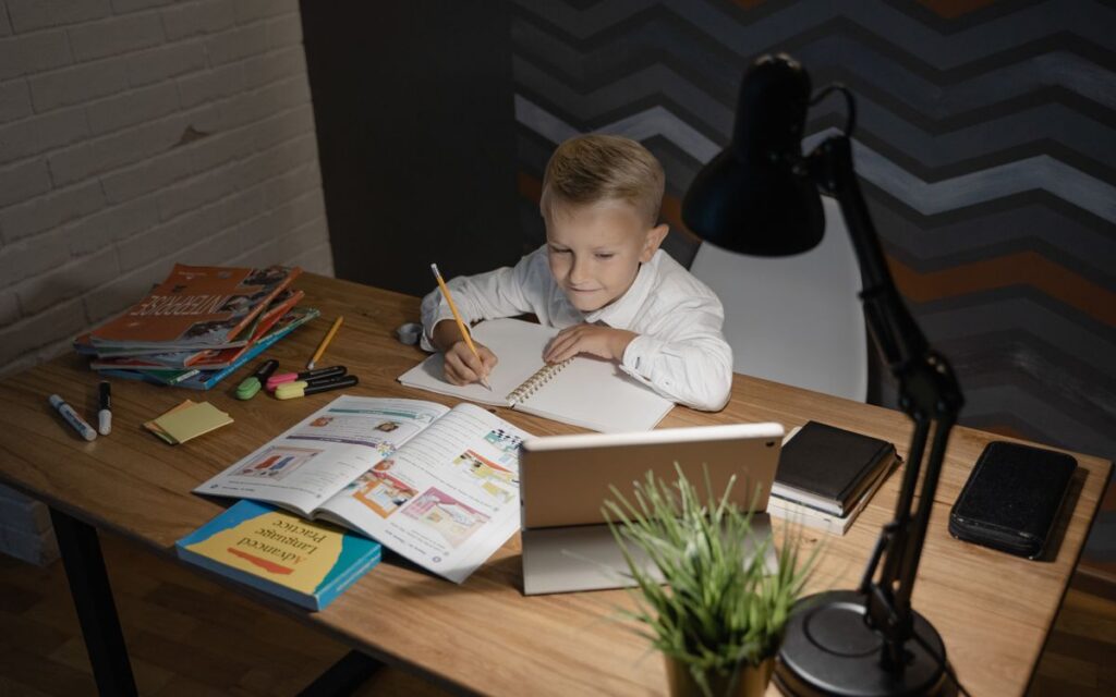 A young boy focused on studying at a wooden table with a tablet and books nearby under a desk lamp.