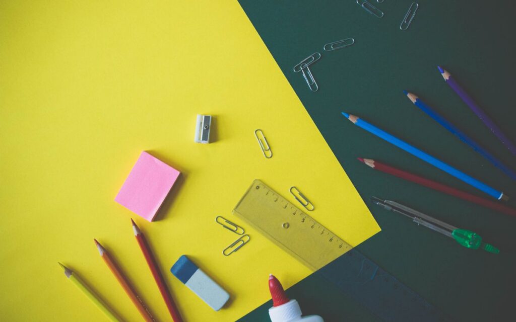 Top view of assorted school supplies like pencils, glue, and paper clips on a yellow and green background.