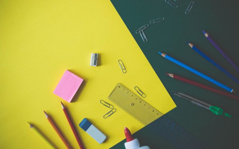 Top view of assorted school supplies like pencils, glue, and paper clips on a yellow and green background.