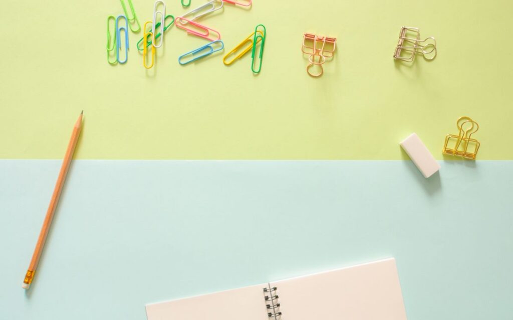 Top view of colorful stationery items, including a notebook, pencil, paperclips, and an eraser on pastel background.