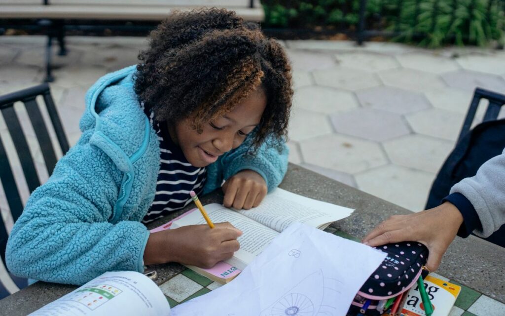 Happy African American child in casual clothes sitting at table with pencil and writing homework while spending time wit