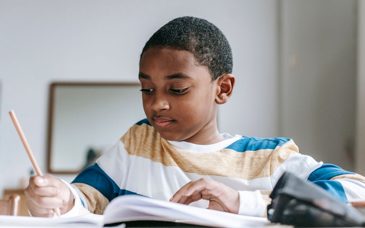 Content African American schoolboy sitting at desk and doing home assignment and writing in notebook