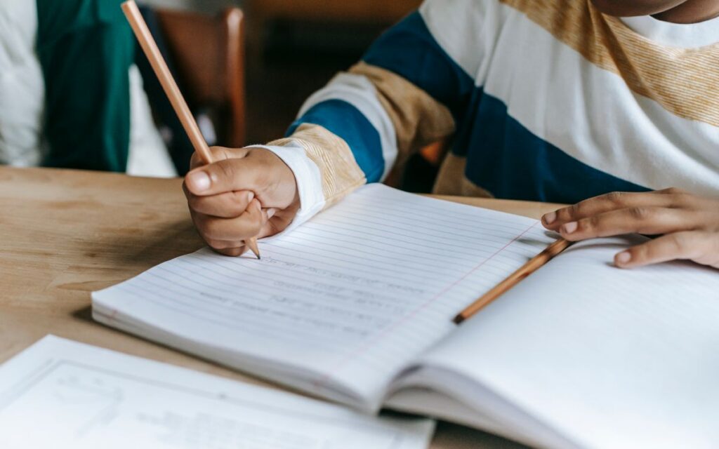 High angle of crop unrecognizable little African American boy writing in notebook while doing schoolwork sitting at tabl