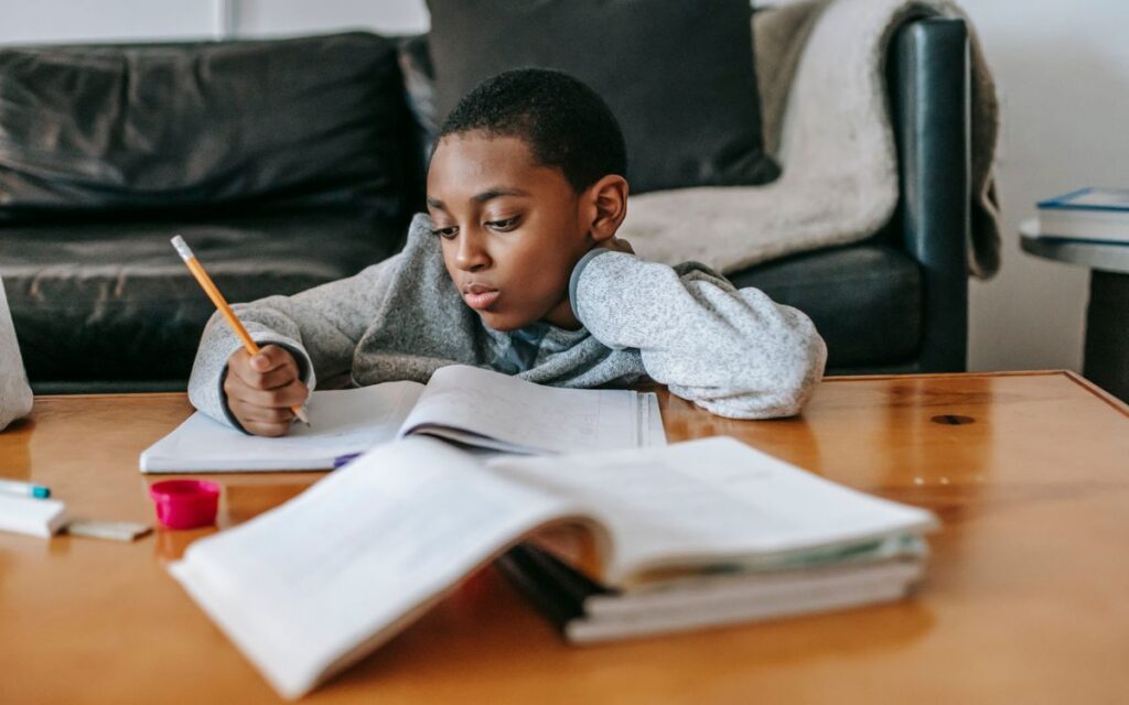 Focused African American boy writing in copybook with pencil while doing homework at wooden table