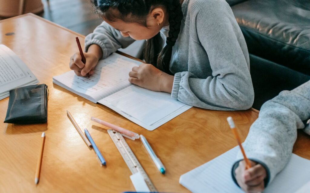 Crop anonymous children sitting at wooden table and writing in copybooks while doing homework together