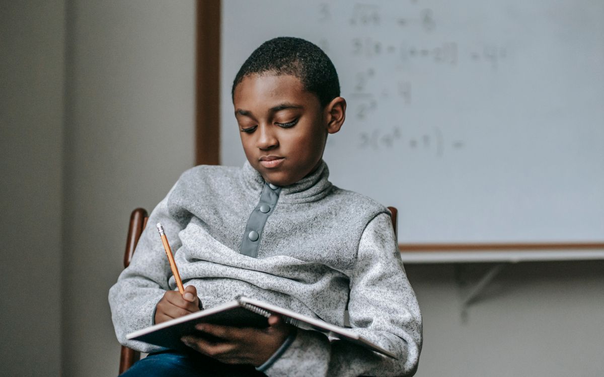 Concentrated African American boy doing tasks in copybook while sitting on wooden chair in classroom