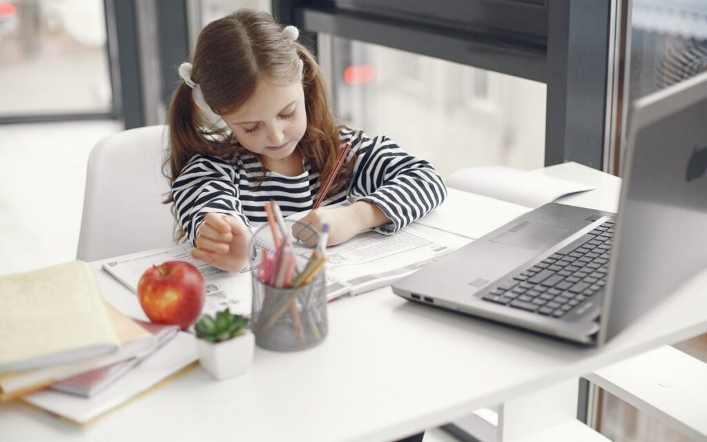 A young girl studies at home using a laptop, engaged with her homework and school supplies.