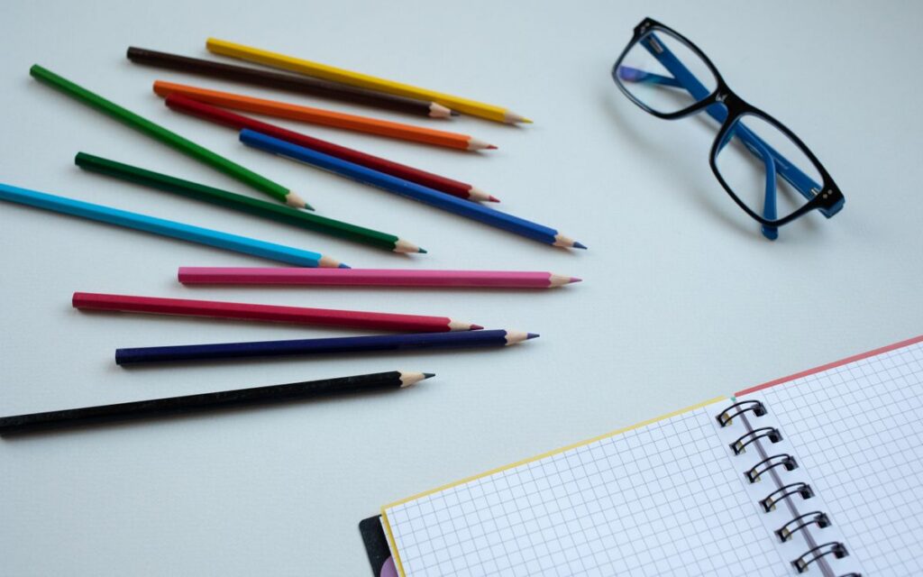 Colorful pencils arranged on a table with a notebook and glasses, ideal for education and creativity themes.
