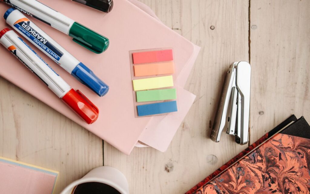 A flat lay of vibrant office supplies including markers, sticky notes, and a stapler on a wooden desk.