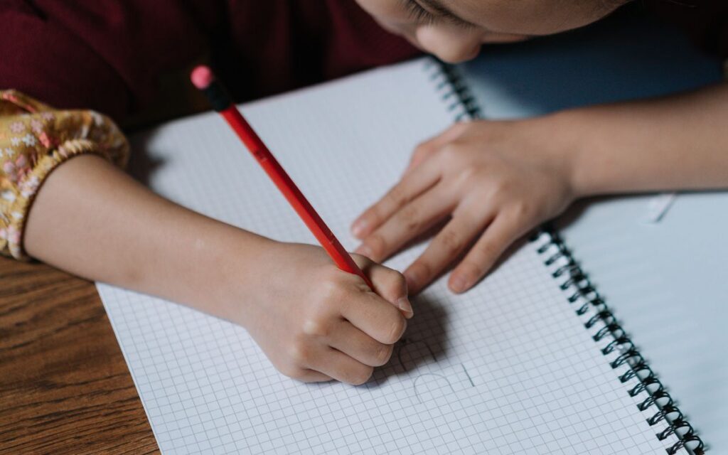 A child writes in a spiral notebook with a red pencil on a wooden desk.